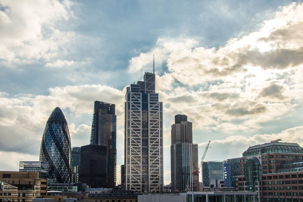 The City of London skyline seen across rooftops, with towers and cranes under a bright, broken cloudscape